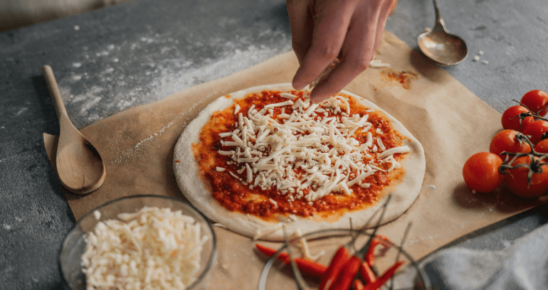 A hand sprinkling cheese onto a raw round pizza on a baking sheet, surrounded by bowls of cheese, chilies, and cherry tomatoes.