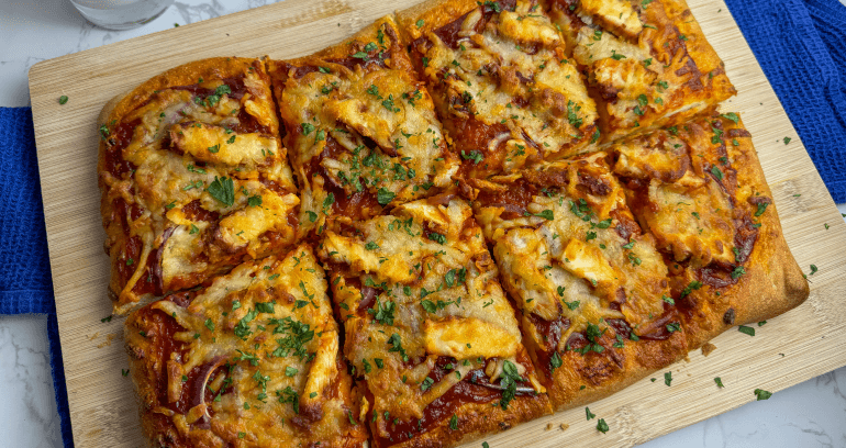 A rectangular pizza topped with chicken, cheese, and barbecue laid on a wooden board on top of a marble surface.