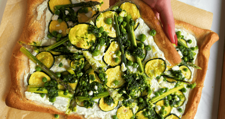 A hand holds a slice of rectangular pizza with feta, ricotta, asparagus, courgette, green peas, and parmesan, with more pizza in the background.