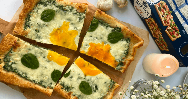 A rectangular pizza topped with eggs, spinach, and basil is cut into 4 pieces on a wooden board by a Jus-Rol Classic Pizza base box.