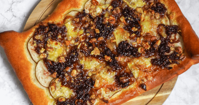 A rectangular pizza lays on a circular wooden board. It’s topped with pear, red onion, brie, honey, and walnuts.