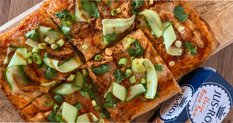 A rectangular pizza on a wooden board topped with chicken, hoisin sauce, mozzarella, cucumber, spring onions, and coriander.