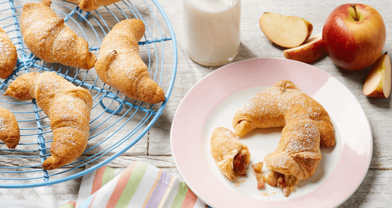 Plate with a sliced croissant filled with fruit, beside whole croissants on a cooling rack, a glass of milk, sliced apples, and a striped cloth on a rustic wooden surface.