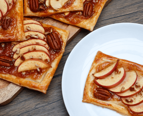 A wooden cutting board displaying fresh caramel, apple and pecan pastry slices