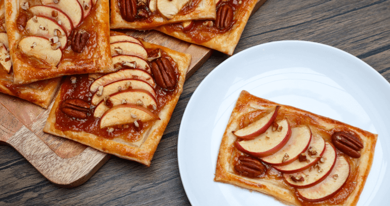 A wooden cutting board displaying fresh caramel, apple and pecan pastry slices