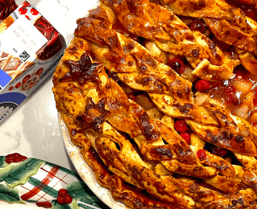 Baked cranberry and pear pie with a lattice crust, a scoop of ice cream on a plate, Jus-Rol pastry packages, a pear, an orange, and a "Cookies for Santa" plate on a marble surface.