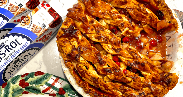 Baked cranberry and pear pie with a lattice crust, a scoop of ice cream on a plate, Jus-Rol pastry packages, a pear, an orange, and a "Cookies for Santa" plate on a marble surface.