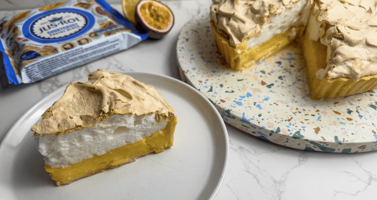 A slice of lemon meringue pie on a plate with the whole pie in the background, next to a package of Jus-Rol shortcrust pastry and half a passion fruit