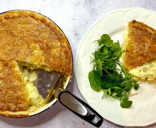 A Three Cheese, Caramelised Onion & Potato Pie served in a circular pie dish next to a plate with a slice of the pie and side salad